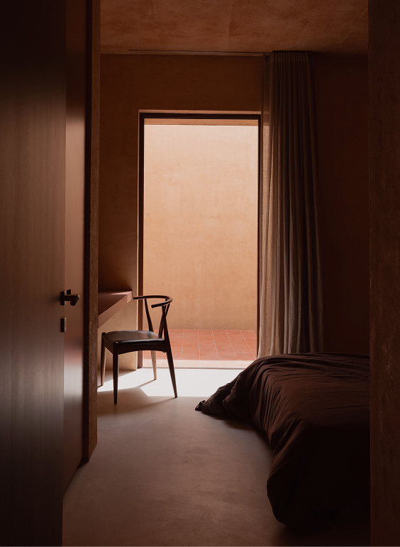 Bedroom looking past a chair toward a doorway opening onto a sunlit terracotta wall